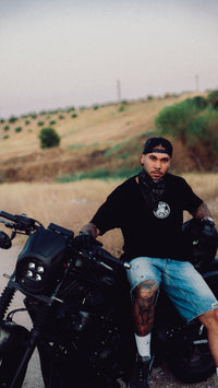 Modelo con la camiseta RAW CUTS apoyado en una moto custom en el campo
